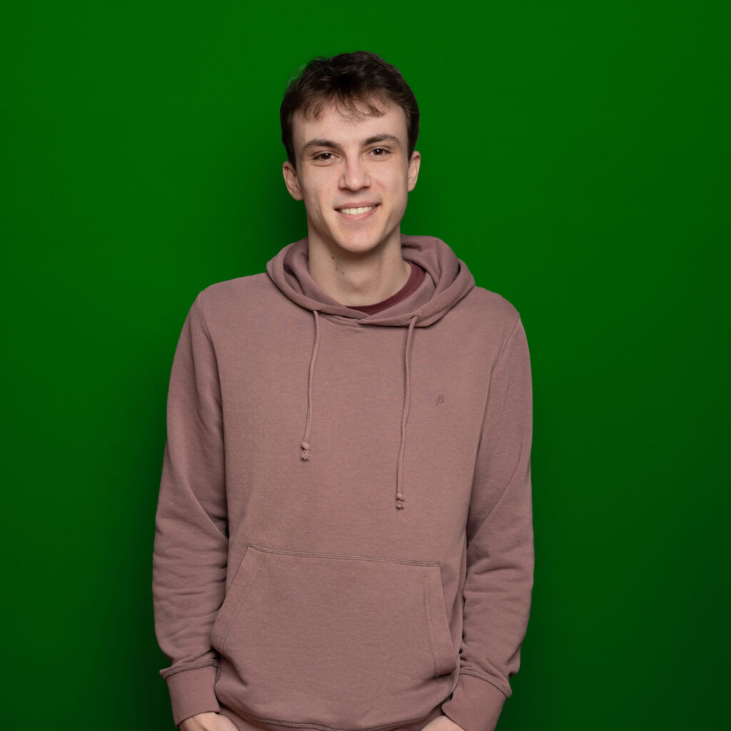 Bild von Luca Moriconi, einem jungen Mann mit heller Haut und kurzen braunen Haaren. Er trägt einen lila-farbenen Pullover und blickt lächelnd in die Kamera.
