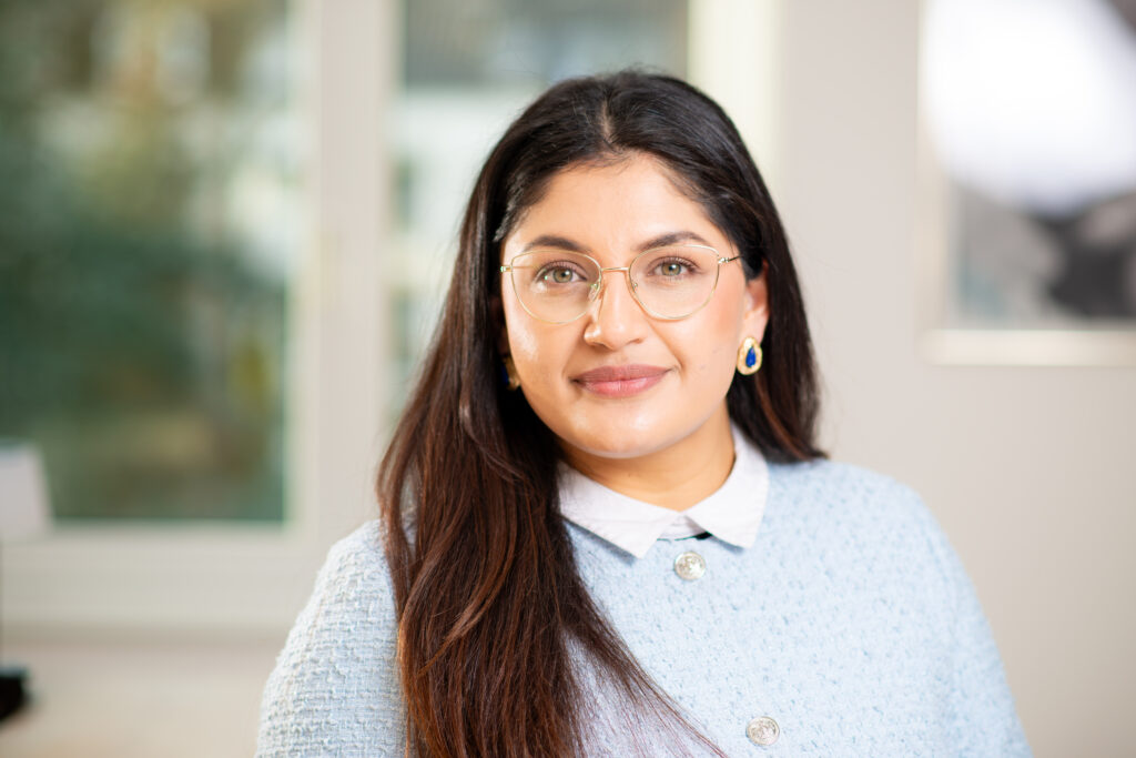 Foto von Mouska Malek, einer jungen Frau mit brauner Haut und dunkelbraunen Haaren. Sie trägt eine silberne Brille und ein hellblauen Strickpullover mit weißem Kragen und blickt lächelnd in die Kamera.