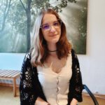 Foto von Leonie Fischer, einer jungen Frau mit heller Haut und schulterlangen braunen Haaren. Sie trägt eine eckige goldene Brille, ein helles Kleid und eine dunkle Strickjacke und blickt lächelnd in die Kamera.