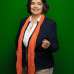 Foto von Dr. Hildegard Bedarff, einer Frau mit heller Haut und kinnlangen, gelockten, schwarz-grauen Haaren. Sie trägt eine weiße Bluse, einen dunkelblauen Blazer und einen orangefarbenen Schal und blickt lächelnd in die Kamera, mit den Händen gestikulierend.