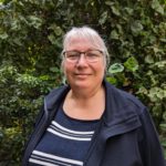 Foto von Andrea Dreffein-Hahn, einer Frau mit heller Haut und grauen Haaren. Sie trägt eine dunkle Brille, ein blau gestreiftes Shirt und eine dunkelblaue Jacke und blickt lächelnd in die Kamera.