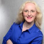 Foto von Anja Schneider, einer Frau mit heller Haut und langen, welligen, blonden Haaren. Sie trägt eine rahmenlose runde Brille und eine blaue Bluse und blickt lächelnd in die Kamera, die Arme locker vor dem Körper verschränkt.