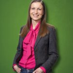 Foto von Anne-Kathrin Ahsbahs, einer Frau mit heller Haut und langen braunen Haaren. Sie trägt eine rosafarbene Bluse und einen grauen Blazer und blickt lächelnd in die Kamera, die Hände locker in den Hostentaschen.
