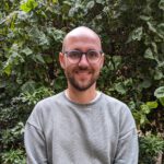 Foto von Arne Heinold, einem Mann mit heller Haut, kurzrasierten Haaren, einem braunen Bart und einer runden grau-blauen Brille. Er trägt einen grauen Pullover und blickt lächelnd in die Kamera. Hinter ihm ein Baum.