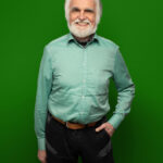Foto von Bent Schubert, einem Mann mit heller Haut, kurzen weißen Haaren und einem weißen Bart. Er trägt ein hellgrünes Hemd und ein gemustertes Halstuch und blickt lächelnd in die Kamera, eine Hand locker in der Hosentasche.