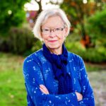Foto von Dagmar Sydow-Graen, einer Frau mit heller Haut und kinnlangen grauen Haaren. Sie trägt eine eckige schwarze Brille, eine blaue Bluse und ein dunkelblaues Halstuch und blickt lächelnd in die Kamera, die Arme locker vor dem Körper verschränkt.