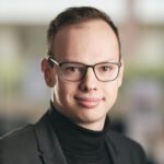 Foto von Florian Juhl, einem jungen Mann mit heller Haut, kurzen braunen Haaren und einer eckigen schwarzen Brille. Er trägt einen schwarzen Rollkragenpullover und ein dunkelgraues Sakko und blickt lächelnd in die Kamera.