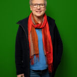 Foto von Ines Strehlau, einer Frau mit heller Haut und kurzen grauen Haaren. Sie trägt eine eckige braune Brille, ein blaues Shirt, eine schwarze Fleecejacke und einen orangefarbenen Schal und blickt lächelnd in die Kamera, eine Hand in der Jackentasche.