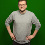 Foto von Jan Mackenthun, einem Mann mit heller Haut, kurzen braunen Haaren und einem kurzen braunen Bart. Er trägt eine dunkle eckige Brille und einen grauen Pullover und blickt lächelnd in die Kamera, die Hände an den Hüften.
