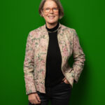 Foto von Lore Möhring, einer Frau mit heller haut und kinnlangen grauen Haaren. Sie trägt eine eckige rote Brille, einen schwarzen Rollkragenpullover und einen geblümten Blazer und blickt lächelnd in die Kamera, eine Hand locker in der Hosentasche.