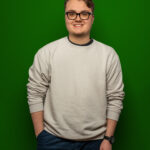 Foto von Lukas Unger, einem jungen Mann mit heller Haut und kurzen braunen Haaren. Er tägt eine eckige schwarze Brille und einen hellgrauen Pullover und blickt lächelnd in die Kamera, eine Hand locker in der Hosentasche.