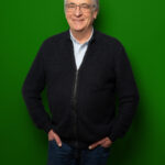 Foto von Mathias Schmitz, einem Mann mit heller Haut und kurzen grauen Haaren. Er trägt eine eckige graue Brille, ein hellblaues Hemd und eine dunkelgraue Strickjacke und blickt lächelnd in die Kamera, die Hände in den Hostentaschen.