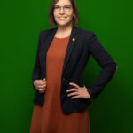 Foto von Nadine Mai, einer Frau mit heller Haut und kinnlangen braunen Haaren. Sie trägt eine runde schwarze Brille, ein dunkelrotes Kleid und einen schwarzen Blazer mit einem kleinen Anstecker in Form einer Sonnenblume am Revers und blickt lächelnd in die Kamera, die Hände an den Hüften.