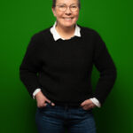 Foto von Regina Flesken, einer Frau mit heller Haut und kurzen grauen Haaren. Sie trägt eine dunkle runde Brille, eine weiße Bluse und einen schwarzen Strickpullover und blickt lächelnd in die Kamera, die Hände locker in den Hosentaschen.