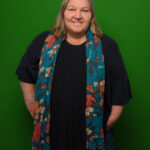 Foto von Sabine Loof, einer Frau mit heller Haut und schulterlangen grau-braunen Haaren. Sie trägt eine schwarze Bluse und einen bunt gemusterten Schal und blickt lächelnd in die Kamera, die Hände an den Hüften.