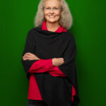 Foto von Sabine Schaefer-Maniezki, einer Frau mit heller Haut und langen, lockigen, grauen Haaren. Sie trägt eine rahmenlose Brille, eine rote Bluse und einen schwarzen Pocho und blickt lächelnd in die Kamera, die Arme vor dem Körper verschränkt.