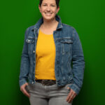 Foto von Sonja Vogt, einer Frau mit heller Haut und kurzen schwarzen Haaren. Sie trägt ein orangefarbenes T-Shirt und eine blaue Jeansjacke und blickt lächelnd in die Kamera, die Daumen locker in den Hosentaschen.