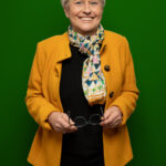 Foto von Susanne von Soden-Stahl, einer Frau mit heller Haut und kurzen grauen Haaren. Sie trägt einen orangefarbenen Blazer und ein buntes Halstuch und blickt lächelnd in die Kamera. In den Händen hält sie ihre dunkle Brille.