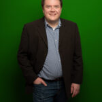 Foto von Sven Herrmann, einem Mann mit heller Haut und kurzen braunen Haaren. Er trägt ein weiß-blau kariertes Hemd und ein offenes schwarzes Sakko und blickt lächelnd in die Kamera, eine Hand locker in der Hosentasche,