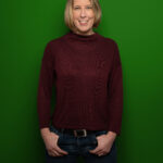 Foto von Sylvia Jacobi, einer Frau mit heller haut und kinnlangen blonden Haaren. Sie trägt einen dunkelroten Rollkragenpullover und blickt lächelnd in die Kamera, die Daumen in den Hosentaschen.