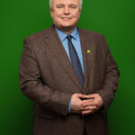 Foto von Thomas Grabau, einem Mann mit heller Haut und kurzen grauen Haaren. Er trägt ein hellblaues Anzughemd, eine dunkelblaue Krawatte und ein braunes Sakko mit einem kleinen Anstecker in Form einer Sonnenblume am Revers und blickt lächelnd in die Kamera, die Hände locker vor dem Körper gefaltet.