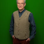 Foto von Thorsten Fischer, einem Mann mit heller Haut und kurzen grauen Haaren. Er trägt eine runde schwarze Brille, ein blaues Jeanshemd und eine graue Weste und blickt lächelnd in die Kamera, die Hände locker in den Hostentaschen.