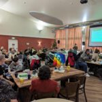 Foto von einer Kreismitgliederversammlung im Lustis in Schenefeld. Es ist weihnachtlich dekoriert und die Mitglieder sitzen an vielen Gruppentischen und hören Lukas Unger zu, der gerade mit dem Mikro in der Hand am Redepult steht.