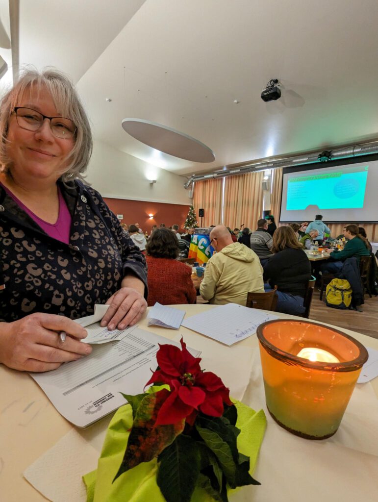 Foto von Andrea Dreffein-Hahn, die an einem Stehtisch vor der Kreismitgliederversammlung die Einlasskontrolle macht und fröhlich in die Kamera lächelt.