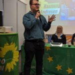 Foto von Lukas Unger bei einer Rede auf einer Kreismitgliederversammlung beim VfL Pinneberg. Er hält das Mikro in der einen Hand und gestikuliert mit der anderen. Hinter ihm das mit einer grünen Europafahne dekorierte Präsidium, an dem Anja Schneider sitzt und zuhört. Neben ihm ein Stehtisch, der mit einer Fahne mit dem Logo von BÜNDNIS 90/DIE GRÜNEN dekoriert ist. Darauf steht ein Beamer, der eine Präsentation an die Wand hinter dem Präsidium wirft. Darauf ist ein Bild und der Namensschriftzug von Europakandidat Rasmus Andresen zu sehen.