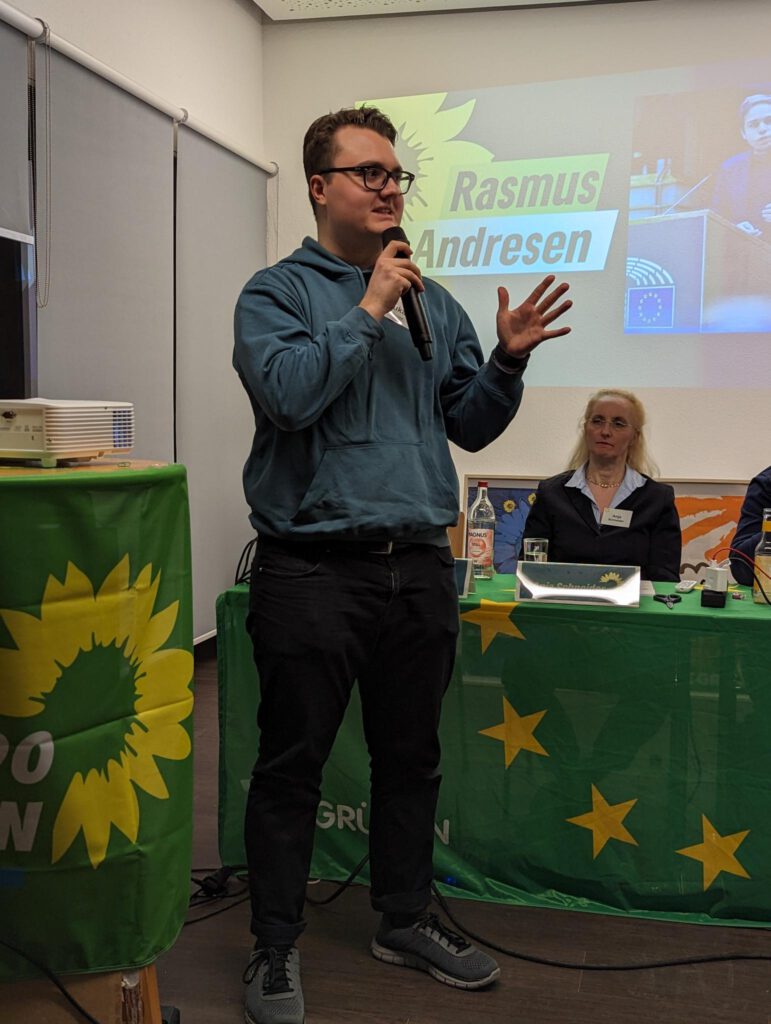 Foto von Lukas Unger bei einer Rede auf einer Kreismitgliederversammlung beim VfL Pinneberg. Er hält das Mikro in der einen Hand und gestikuliert mit der anderen. Hinter ihm das mit einer grünen Europafahne dekorierte Präsidium, an dem Anja Schneider sitzt und zuhört. Neben ihm ein Stehtisch, der mit einer Fahne mit dem Logo von BÜNDNIS 90/DIE GRÜNEN dekoriert ist. Darauf steht ein Beamer, der eine Präsentation an die Wand hinter dem Präsidium wirft. Darauf ist ein Bild und der Namensschriftzug von Europakandidat Rasmus Andresen zu sehen.