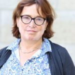 Foto von Eka von Kalben, einer Frau mit heller Haut mit Vitiligo und kinnlangen braunen Haaren. Sie trägt eine dunkelbraune Brille und eine blaue gemusterte Bluse und blickt lächelnd in die Kamera.