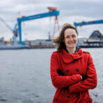 Foto von Ingrid Nestle, einer Frau mit heller Haut und kinnlangen braunen Haaren. Sie trägt einen roten Trenchcoat, schwarze Handschuhe und blickt lächelnd in die Kamera, die Hände vor dem Körper verschränkt. Im Hintergrund ist ein Frachthafen zu sehen.