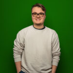 Foto von Lukas Unger, einem jungen Mann mit heller Haut und kurzen braunen Haaren. Er tägt eine eckige schwarze Brille und einen hellgrauen Pullover und blickt lächelnd in die Kamera, eine Hand locker in der Hosentasche.