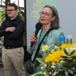 Sylvia und Lukas Unger stehen auf der Bühne. Sylvia spricht ins Mikrofon. Im Vordergrund ein gelber Blumenstrauß auf einem Tisch.