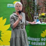 Foto von Susanne von Soden-Stahl, einer Frau mit heller Haut und kurzen weißen Haaren. Sie trägt eine runde Brille und ein olivfarbenes Kleid und hält eine Rede, neben einem Stehtisch stehend, der mit einer grünen Fahne und einem Blumenstrauß dekoriert ist.