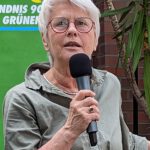Foto von Susanne von Soden-Stahl, einer Frau mit heller Haut und kurzen weißen Haaren. Sie trägt eine runde Brille und ein olivfarbenes Kleid und hält eine Rede, neben einem Stehtisch stehend, der mit einer grünen Fahne und einem Blumenstrauß dekoriert ist.