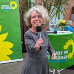 Foto von Katrin Langensiepen, einer Frau mit heller Haut, lockigen grau-braunen Haaren und einer sichtbaren körperlichen Behinderung, durch die sie sehr kurze Arme hat. Sie trägt eine eckige Brille und ein kariertes Kleid und spricht ausdrucksstark mit fröhlichem Blick in ein Mikrofon. Im Hintergrund sieht man eine grüne Stellwand mit der Aufschrift "BÜNDNIS 90/DIE GRÜNEN" und einen mit einer grünen Fahne und einem Blumenstrauß dekorierten Tisch.
