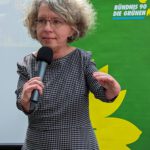 Foto von Katrin Langensiepen, einer Frau mit heller Haut, lockigen grau-braunen Haaren und einer sichtbaren körperlichen Behinderung, durch die sie sehr kurze Arme hat. Sie trägt eine eckige Brille und ein kariertes Kleid und spricht ausdrucksstark mit ernstem Blick in ein Mikrofon. Im Hintergrund sieht man eine grüne Stellwand mit der Aufschrift "Bündnis 90 Die Grünen".