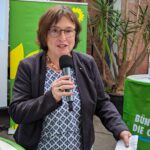 Eka von Kalben, eine Frau mit heller Haut und kurzen braunen Haare, hält eine Rede. Sie trägt eine runde Brille und eine gemusterte Bluse mit einem dunkelblauen Blazer und spricht fröhlich ins Mikrofon.