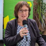 Eka von Kalben, eine Frau mit heller Haut und kurzen braunen Haare, hält eine Rede. Sie trägt eine runde Brille und eine gemusterte Bluse mit einem dunkelblauen Blazer und spricht fröhlich ins Mikrofon.