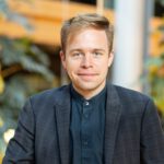 Foto von Rasmus Andresen, einem Mann mit heller Haut und kurzen blonden Haaren. Er trägt ein dunkelblaues Hemd und ein offenes dunkelblaues Sakko und blickt lächelnd in die Kamera.