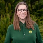 Foto von Ann Christin Hahn, einer Frau mit heller Haut und langen braunen Haaren. Sie trägt eine runde schwarze Brille und einen dunkelgrünen Pullover mit einer Sonnenblume und blickt lächelnd in die Kamera.