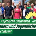 Gruppenfoto von Grünen und Krankenhausmitarbeitenden vor der Kinder- und Jugendpsychiatrie in Elmshorn. Darauf der Text: "Psychische Gesundheit von Kindern und Jugendlichen schützen!" Daneben die Sonnenblume der Grünen.