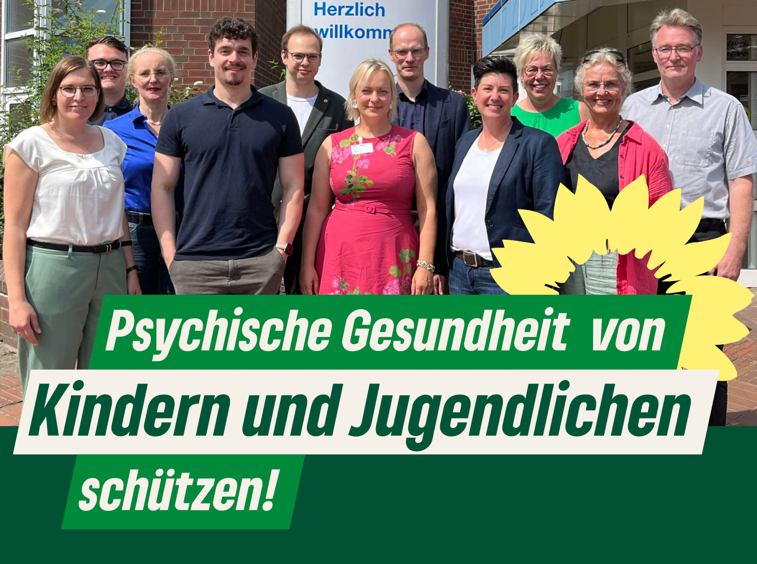 Gruppenfoto von Grünen und Krankenhausmitarbeitenden vor der Kinder- und Jugendpsychiatrie in Elmshorn. Darauf der Text: "Psychische Gesundheit von Kindern und Jugendlichen schützen!" Daneben die Sonnenblume der Grünen.