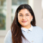 Foto von Mouska Malek, einer jungen Frau mit brauner Haut und dunkelbraunen Haaren. Sie trägt eine silberne Brille und ein hellblauen Strickpullover mit weißem Kragen und blickt lächelnd in die Kamera.