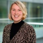 Foto von Denise Loop, einer Frau mit heller Haut und kinnlangen blonden Haaren. Sie trägt eine braune geblümte Bluse und blickt lächelnd in die Kamera.