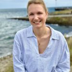 Foto von Marlene Renner, einer jungen Frau mit heller Haut und zu einem Zopf gebundenen blonden Haaren. Sie trägt eine weiß-blau gestreifte Bluse und eine goldene Halskette und blickt fröhlich lächelnd in die Kamera. Im Hintergrund sieht man eine grasbewachsene Küstenlinie und das Meer.