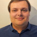 Foto von Sören Lerke, einem jungen Mann mit heller Haut und kurzen braunen Haaren. Er trägt ein dunkelblaues Hemd und blickt freundlich lächelnd in die Kamera. Hinter ihm eine weiße Wand.